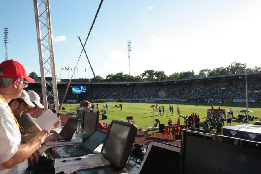Stanowisko dziennikarzy sportowych z laptopami na trybunach stadionu lekkoatletycznego podczas zawodów, z widokiem na murawę i kibiców.