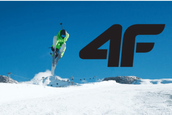 Narciarz w zielonym kombinezonie wykonuje skok na śnieżnym stoku z dużym logo 4F na tle nieba.