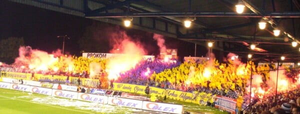 Kibice Arki Gdynia na stadionie, żółto-niebieskie flagi i race podczas meczu piłkarskiego