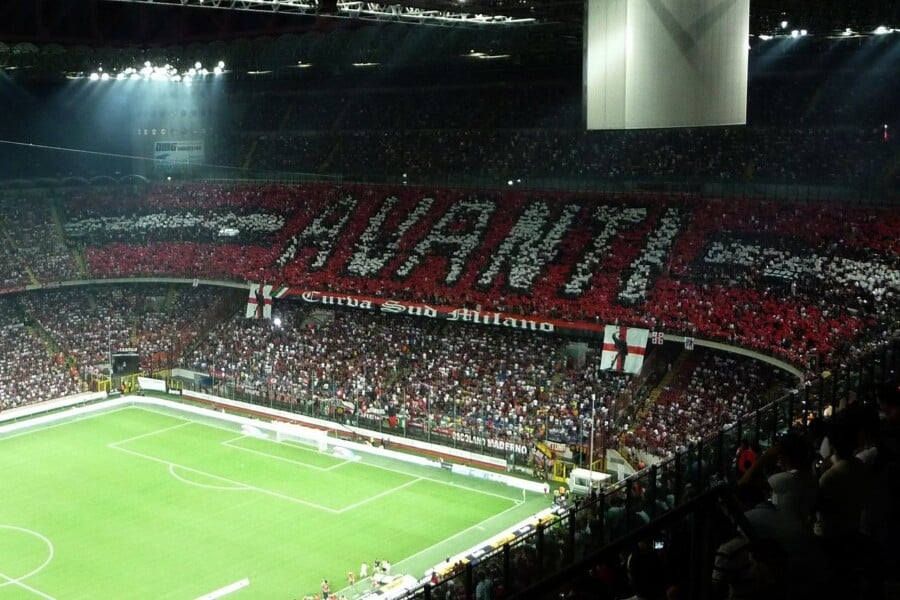 Widok z trybun stadionu San Siro w Mediolanie podczas derbów AC Milan – Inter, z sektorów Curva Sud tworzących choreografię z napisem „AVANTI” nad murawą boiska.