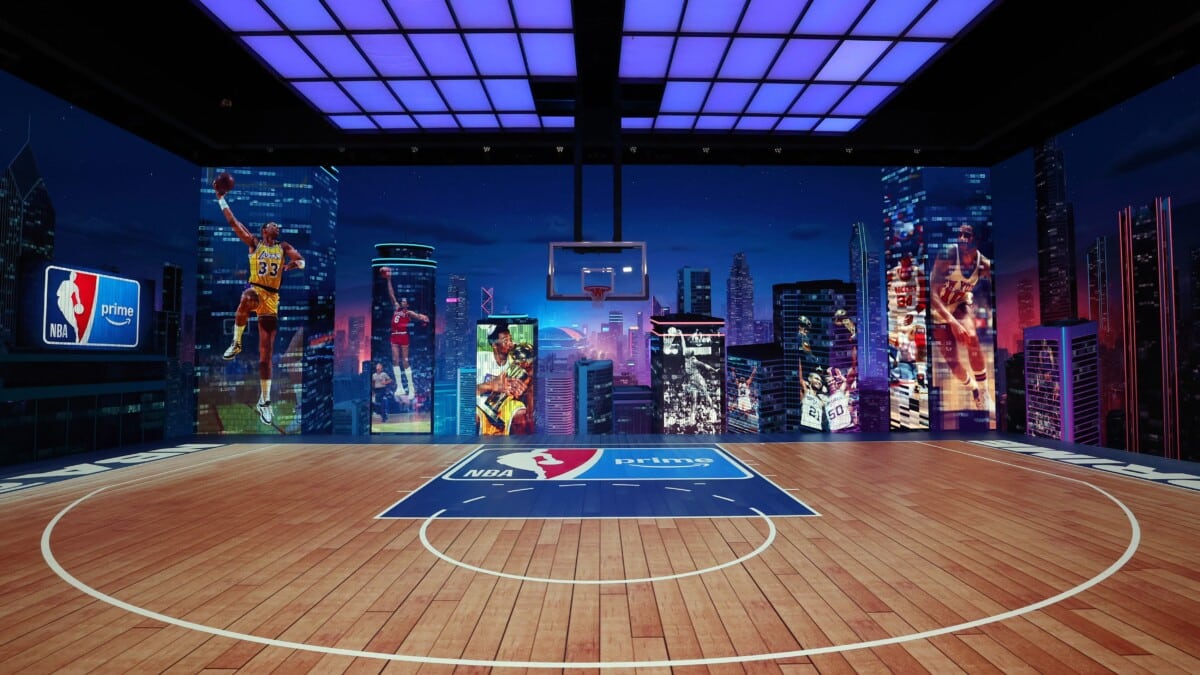 Nowoczesne boisko do koszykówki z logotypami NBA i Prime Video oraz muralami legendarnych graczy NBA na tle panoramy miasta