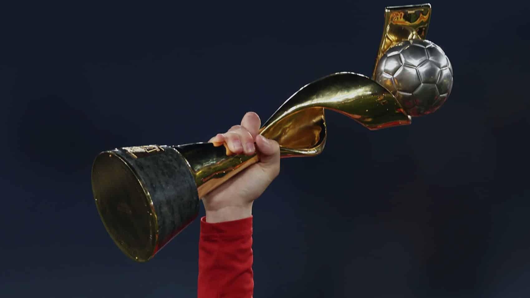 Ręka piłkarki w czerwonej koszulce unosi w górę złoty puchar mistrzostw świata kobiet FIFA z piłką na szczycie na ciemnym tle