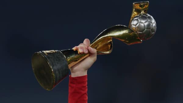 Ręka piłkarki w czerwonej koszulce unosi w górę złoty puchar mistrzostw świata kobiet FIFA z piłką na szczycie na ciemnym tle