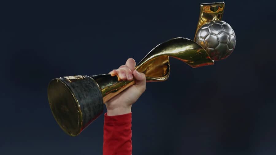 Ręka piłkarki w czerwonej koszulce unosi w górę złoty puchar mistrzostw świata kobiet FIFA z piłką na szczycie na ciemnym tle