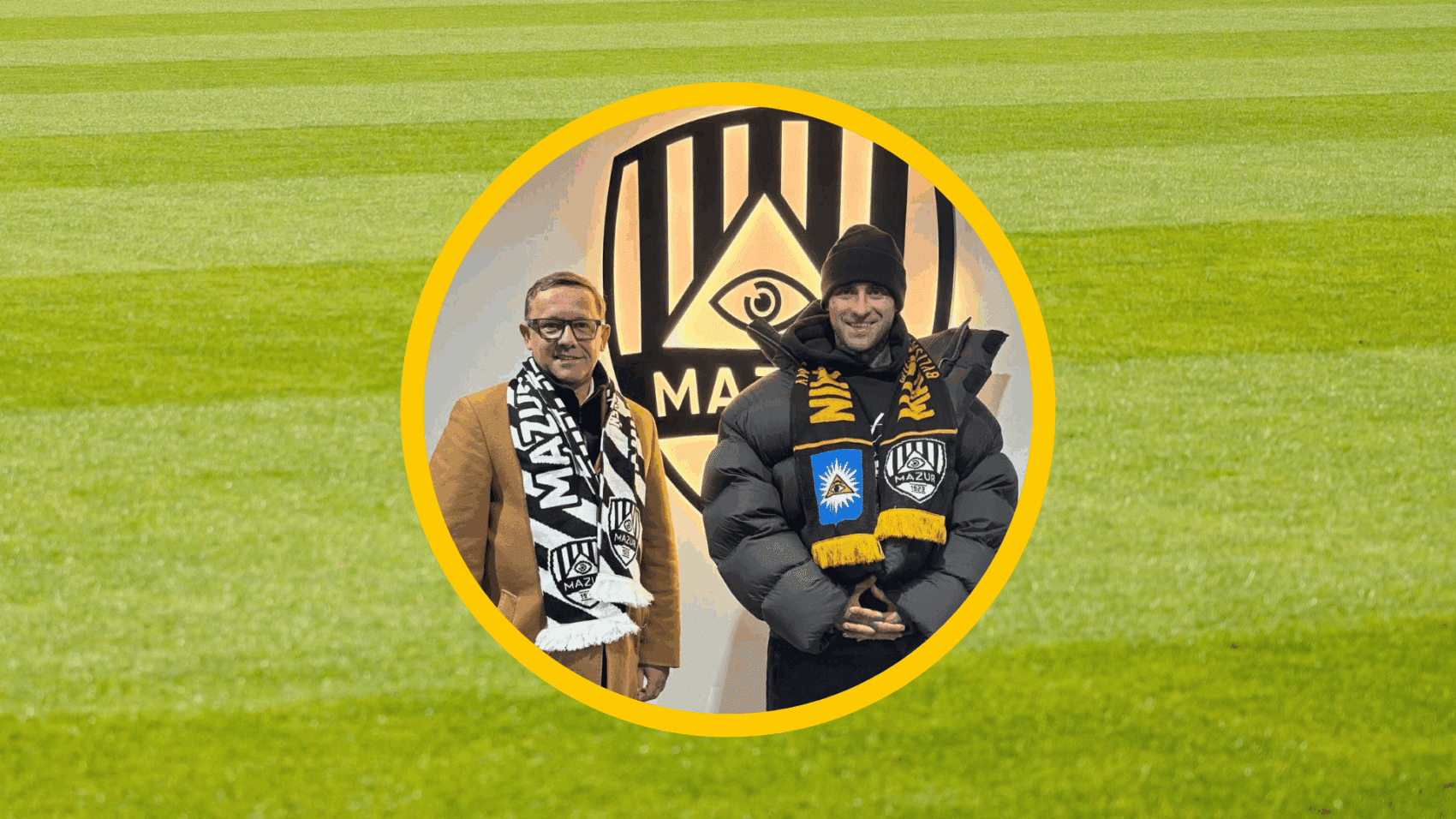 Dwaj mężczyźni w szalikach Mazura Radzymin pozują na tle herbu klubu, symbolizując ogłoszenie nowego dyrektora sportowego.
