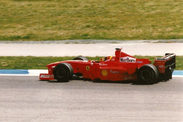 Klasyczny czerwony bolid Ferrari Formuły 1 z lat 90. z dużymi logotypami Marlboro na nosie i Shell na silniku, niebiesko-białe skrzydło tylne, kierowca w kasku, pokonuje zakręt na asfalcie z białą linią i zieloną trawą.