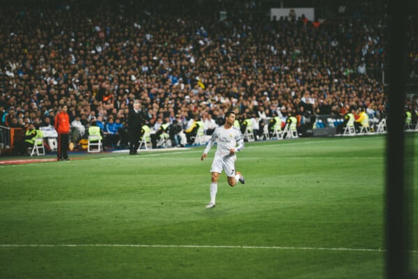 Piłkarz Real Madrid w białej koszulce domowej z krótkim rękawem dynamicznie biegnie po boisku podczas meczu; rozmyte pełne trybuny kibiców w białych barwach, zielona murawa, barierki i krzeseła ochroniarzy w tle.