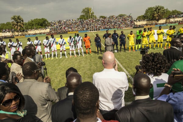 Piłkarze dwóch afrykańskich reprezentacji ustawieni w rzędzie na murawie przed meczem, przy linii bocznej stoją sędziowie i oficjele, a w tle widać wypełnione kibicami trybuny stadionu.