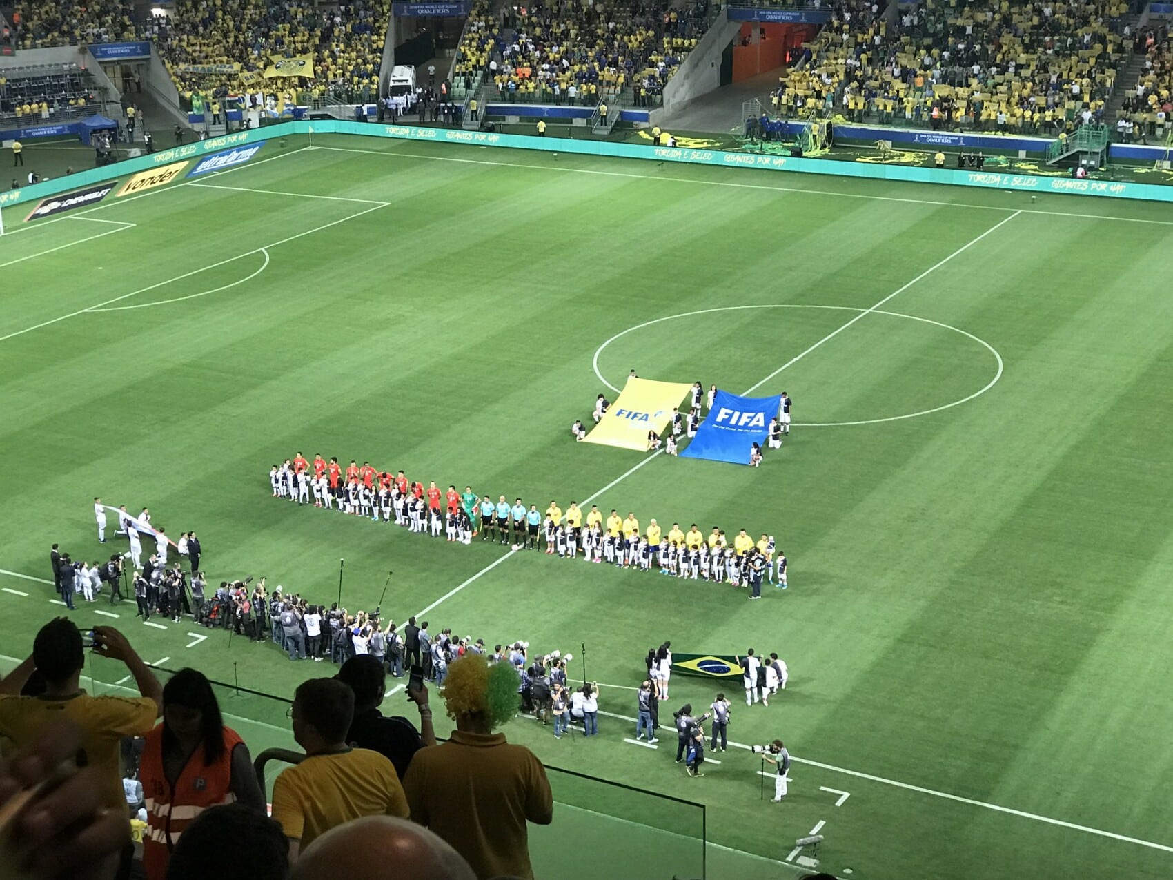 Piłkarze ustawieni w dwóch szeregach przed rozpoczęciem meczu piłki nożnej FIFA na zielonej murawie dużego stadionu, z flagami FIFA na środku boiska i kibicami w żółtych koszulkach na trybunach.