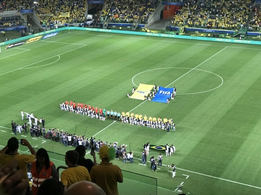 Piłkarze ustawieni w dwóch szeregach przed rozpoczęciem meczu piłki nożnej FIFA na zielonej murawie dużego stadionu, z flagami FIFA na środku boiska i kibicami w żółtych koszulkach na trybunach.