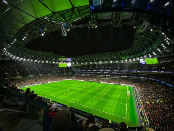 Pełny stadion Tottenham Hotspur Stadium podczas wieczornego meczu Tottenhamu z Liverpoolem w Premier League, z zieloną murawą, kibicami i oświetleniem.