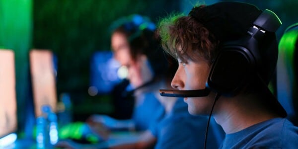 Młoda osoba w słuchawkach z mikrofonem siedzi przy komputerze podczas drużynowej rozgrywki e‑sportowej, w tle widać innych graczy przy monitorach.