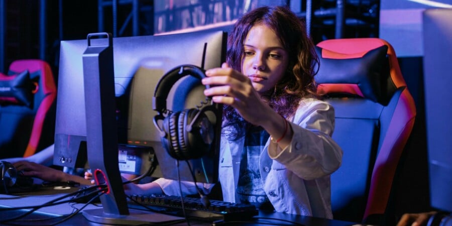 Dziecko siedzi przy biurku z monitorem i kolorowym fotelem gamingowym, trzymając w ręku słuchawki przed rozpoczęciem rozgrywki e‑sportowej na komputerze.