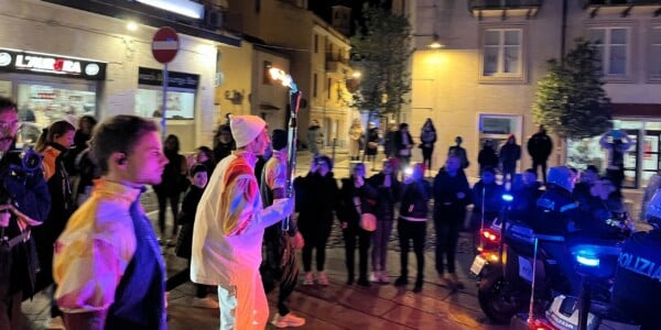 Podczas 8. etapu sztafety znicza olimpijskiego Zimowych Igrzysk Milano Cortina 2026, mężczyzna w białej czapce i wielobarwnym stroju trzyma zapaloną pochodnię; wokół tłum mieszkańców, policja z niebieskimi światłami, nocne ulice Nuoro z budynkami i latarniami.