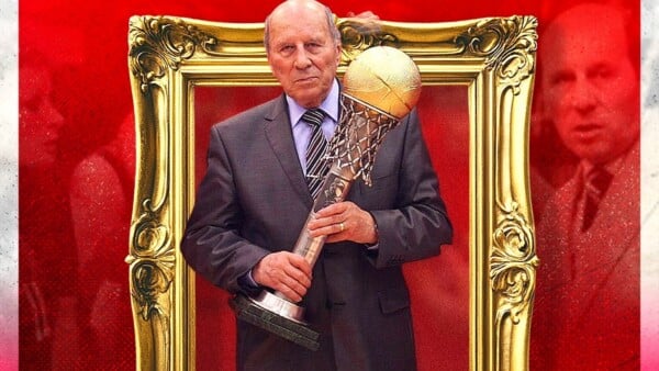 Polski działacz koszykówki w garniturze trzyma dużą, złoto‑srebrną statuetkę w kształcie kosza na tle dekoracyjnej złotej ramy i czerwonego tła.