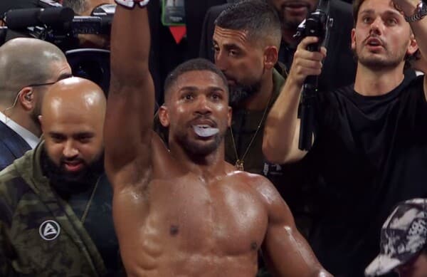 Anthony Joshua (w czarnym topie i łańcuchu) z uniesioną prawą pięścią celebruje wygraną przez KO z Jake'iem Paulem na ringu; wokół niego entuzjastyczny entourage z kamerami i mikrofonami, grudzień 2025, boks heavyweight.