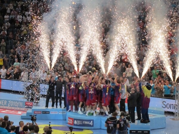 Zespół piłkarzy ręcznych świętuje na podium zdobycie trofeum EHF Champions League, otoczony konfetti i fajerwerkami, przy pełnych trybunach kibiców w hali sportowej.
