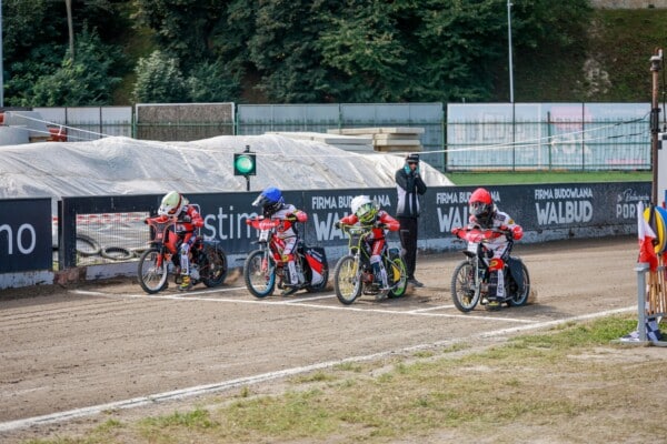 Start wyścigu juniorów na torze żużlowym. Czterech młodych zawodników w pełnym ekwipunku czeka na zielone światło. Tak wyglądają zawody młodzieżowe.