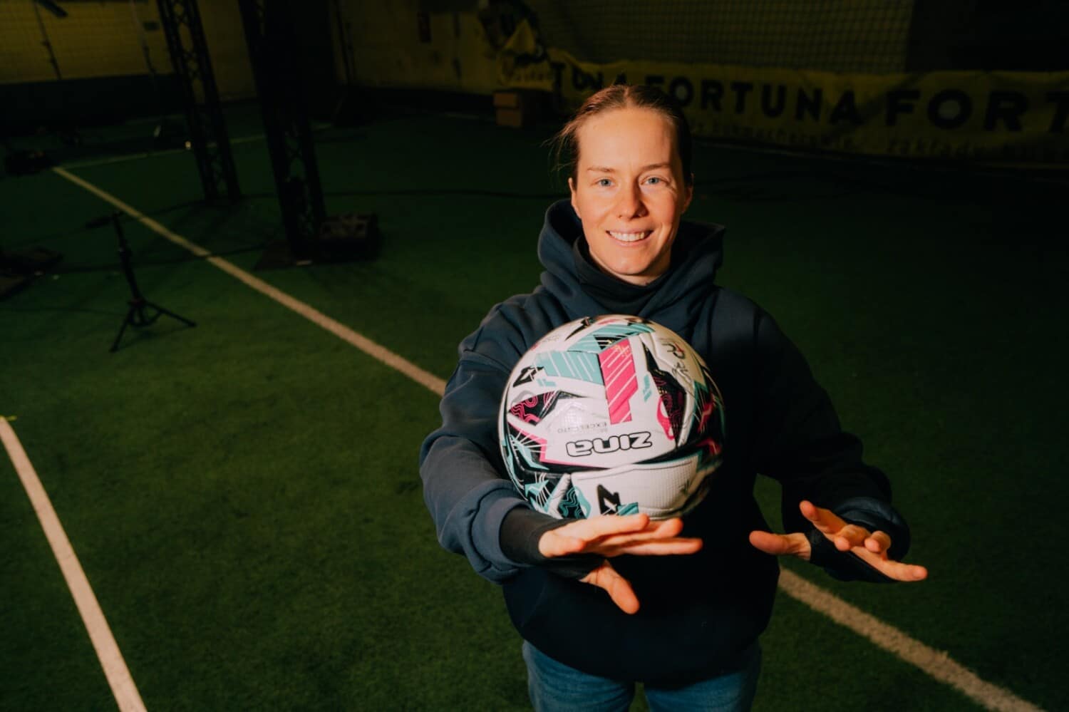 Sabina „Starka” Worosz uśmiecha się do aparatu, trzymając przed sobą białą piłkę do piłki nożnej z kolorowymi wzorami na tle zielonego boiska treningowego.