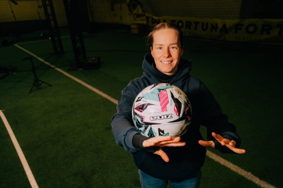 Sabina „Starka” Worosz uśmiecha się do aparatu, trzymając przed sobą białą piłkę do piłki nożnej z kolorowymi wzorami na tle zielonego boiska treningowego.