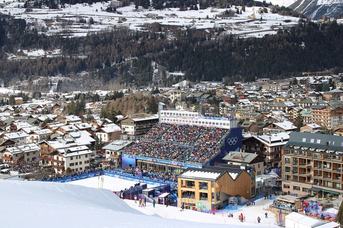 Widok z lotu ptaka na trasę Stelvio w Bormio Stelvio Ski Centre podczas MŚ w narciarstwie alpejskim FIS 2026, zatłoczony stadion z pierścieniami olimpijskimi i panorama zaśnieżonych włoskich Alp.