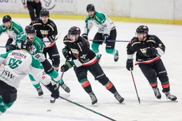 Intensywna akcja podczas meczu hokejowego PHL pomiędzy JKH GKS Jastrzębie w czarno-czerwonych barwach a GKS Tychy w zielono-białych koszulkach; zawodnicy walczą o krążek na lodowisku, Jastrzębianie w natarciu.