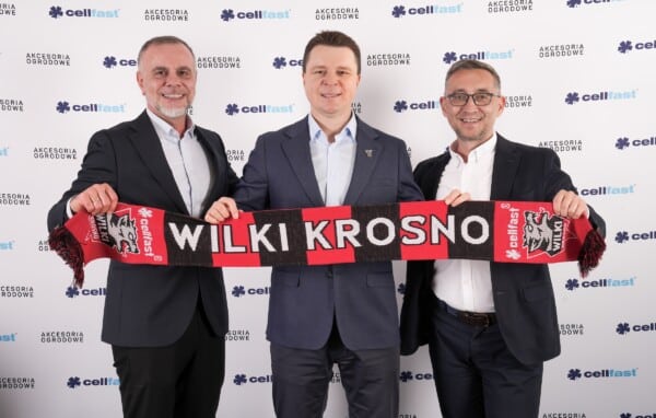 Trzej mężczyźni w garniturach pozują z szarfą "Wilki Krosno" na tle banerów Cellfast na wydarzeniu sponsorskim żużlowego klubu Cellfast Wilki Krosno