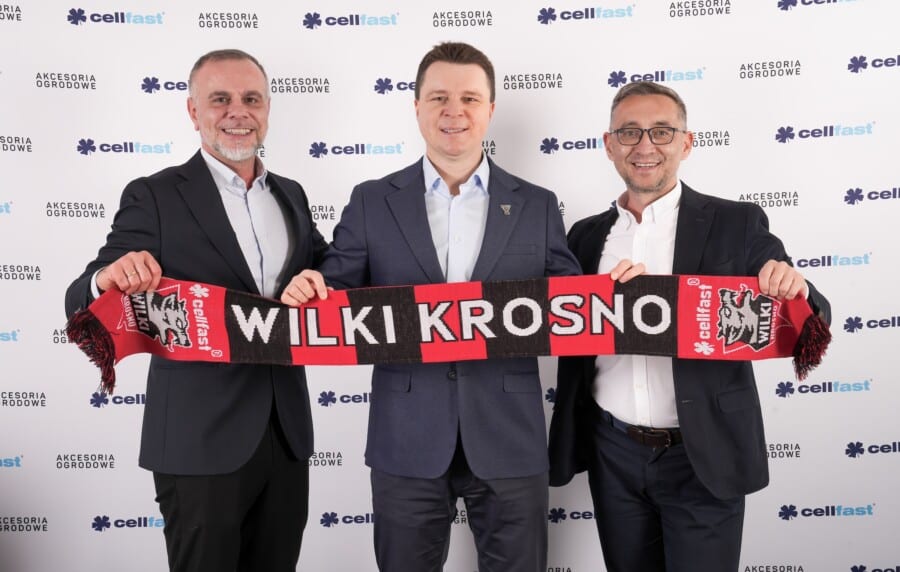Trzej mężczyźni w garniturach pozują z szarfą "Wilki Krosno" na tle banerów Cellfast na wydarzeniu sponsorskim żużlowego klubu Cellfast Wilki Krosno