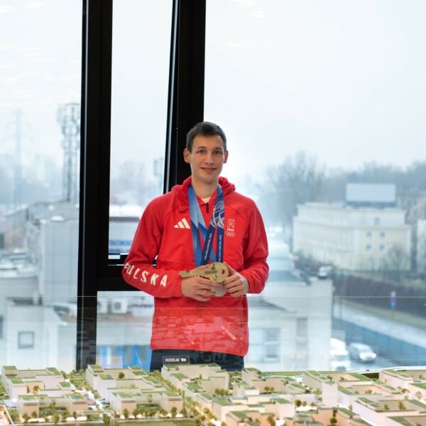 Kacper Tomasiak, polski skoczek narciarski, pozuje w czerwonej bluzie Adidas z medalami zdobytymi na Zimowych Igrzyskach Olimpijskich w Mediolanie-Cortinie 2026, trzymając je przed modelem miasta z widokiem na Warszawę.