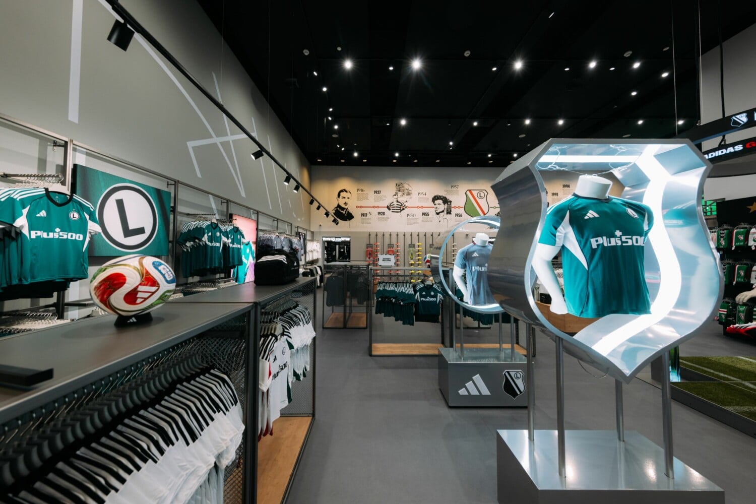 Nowoczesny oficjalny sklep kibica Legia Warszawa z zielonymi koszulkami Adidas, piłką nożną, trofeami i oświetleniem LED – wnętrze pełne gadżetów legionistów przy Łazienkowskiej.