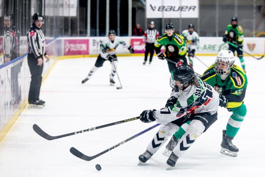Dwie zawodniczki profesjonalnej ligi hokejowej rywalizują na lodowisku. W tle widać sędzię oraz resztę zawodniczek z rywalizujących zespołów