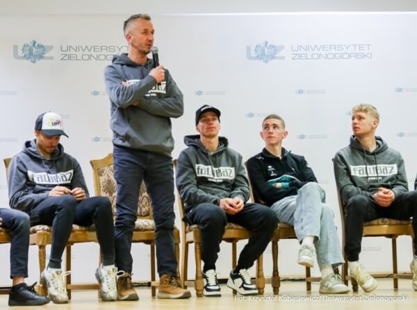 Prezes Stelmet Falubaz Zielona Góra Adam Goliński w szarym bluzie z mikrofonem przemawia do młodych żużlowców w bluzach Fallz na spotkaniu z studentami Uniwersytetu Zielonogórskiego – podpisanie listu intencyjnego o badaniach AI i optymalizacji wyników na torze 4 marca 2026.
