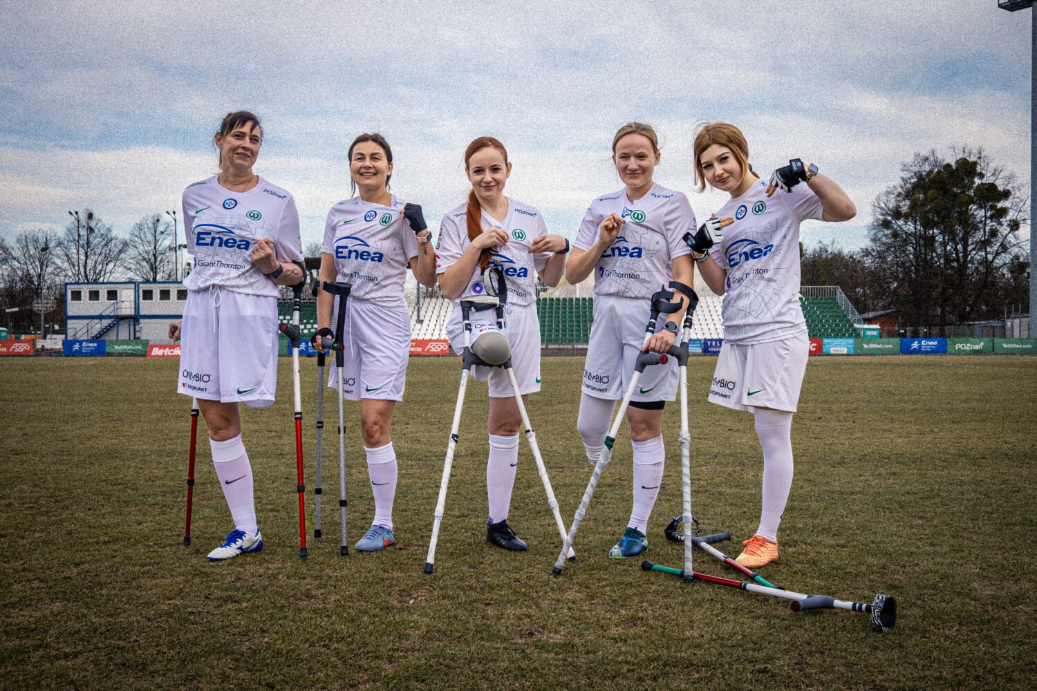 Zawodniczki sekcji Amp Futbolu kobiet Warty Poznań w białych koszulkach z logo Enea stoją na zielonym boisku stadionu, trzymając kule i piłkę amp futbolu; uśmiechnięte pięć piłkarek z niepełnosprawnościami ruchowymi, pochmurne niebo, trybuny w tle.