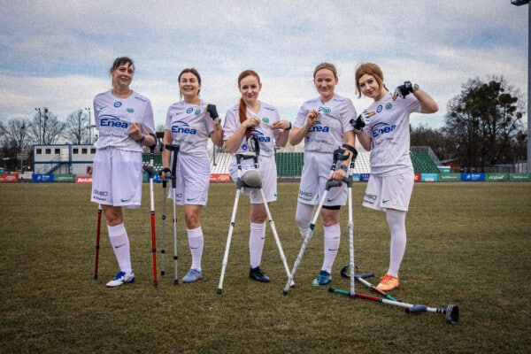 Zawodniczki sekcji Amp Futbolu kobiet Warty Poznań w białych koszulkach z logo Enea stoją na zielonym boisku stadionu, trzymając kule i piłkę amp futbolu; uśmiechnięte pięć piłkarek z niepełnosprawnościami ruchowymi, pochmurne niebo, trybuny w tle.