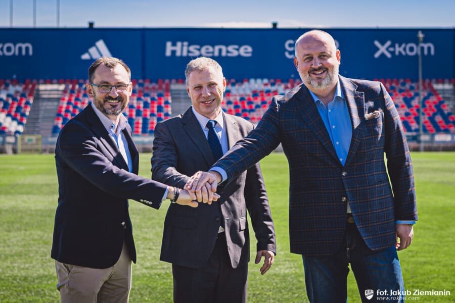 Trzej mężczyźni w garniturach, reprezentujący Raków Częstochowa i Politechnikę Częstochowską, kładą dłonie na sobie w geście porozumienia i współpracy na tle trybun stadionu piłkarskiego.