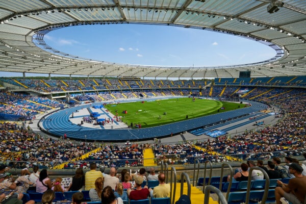 Szeroka panorama wnętrza Stadionu Śląskiego w Chorzowie z perspektywy górnych trybun. Na pierwszym planie widoczni kibice, w głębi niebieska bieżnia lekkoatletyczna otaczająca zieloną murawę oraz charakterystyczne zadaszenie obiektu w słoneczny dzień.