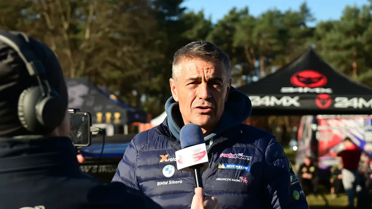 Krzysztof Hołowczyc udziela wywiadu. Stoi w puchowej kurtce przed dziennikarzem, który wystawia mu mikrofon