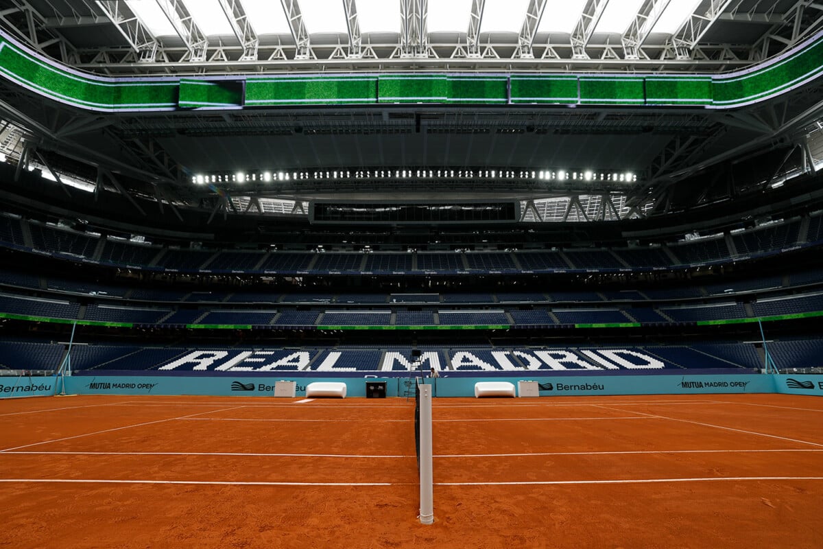 Widok na ceglany kort tenisowy zainstalowany na murawie stadionu Santiago Bernabéu w Madrycie z widocznym napisem Real Madrid na trybunach.