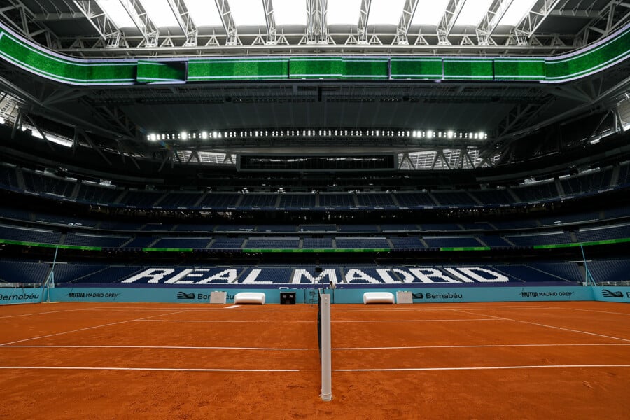 Widok na ceglany kort tenisowy zainstalowany na murawie stadionu Santiago Bernabéu w Madrycie z widocznym napisem Real Madrid na trybunach.