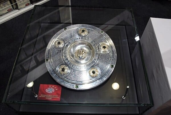 Srebrna patera mistrza Niemiec (Meisterschale Bundesligi) z zielonymi kamieniami szlachetnymi, wyeksponowana w szklanej gablocie z czerwoną tabliczką Bayernu Monachium.