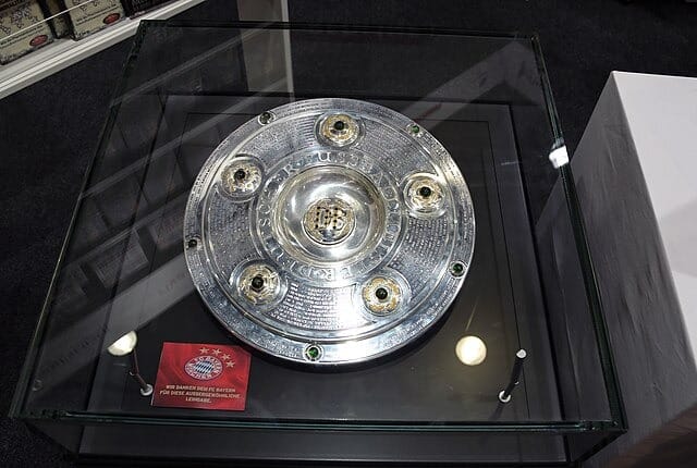 Srebrna patera mistrza Niemiec (Meisterschale Bundesligi) z zielonymi kamieniami szlachetnymi, wyeksponowana w szklanej gablocie z czerwoną tabliczką Bayernu Monachium.