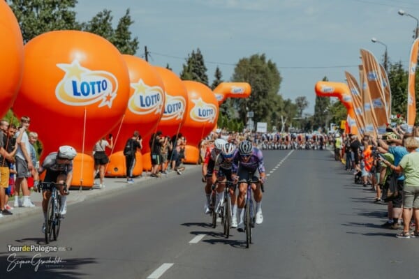 Zbliżenie na czołową grupę kolarzy rywalizujących w Tour de Pologne, przejeżdżających obok rzędu dużych, pomarańczowych balonów reklamowych z logo LOTTO na prostej drodze asfaltowej. W tle kibice i peleton. Wyścig UCI WorldTour w Polsce.