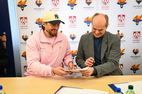 Rektor AWF Katowice prof. dr hab. n. med. Andrzej Małecki w szarym swetrze i Ołeksandr Usyk w różowym polarze z czapką New York Yankees podpisują umowę o współpracy przy banerze AWF Katowice, badanie urazów mózgu w boksie