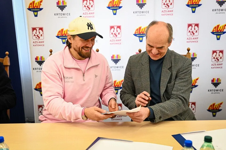 Rektor AWF Katowice prof. dr hab. n. med. Andrzej Małecki w szarym swetrze i Ołeksandr Usyk w różowym polarze z czapką New York Yankees podpisują umowę o współpracy przy banerze AWF Katowice, badanie urazów mózgu w boksie