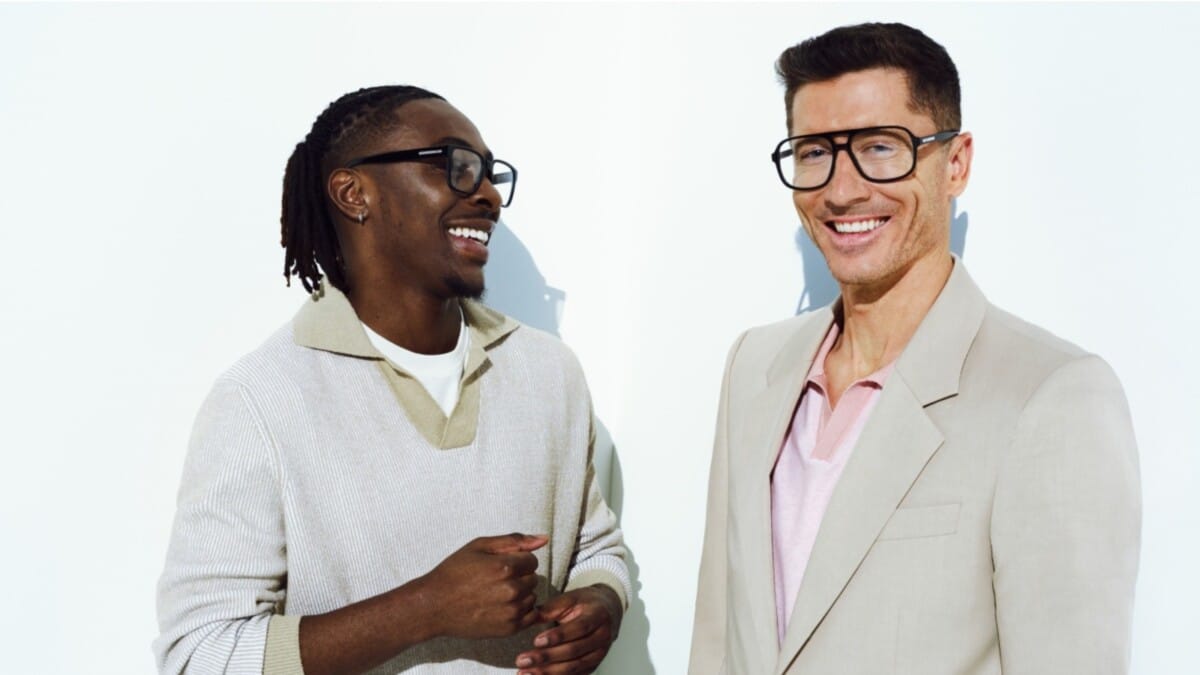 Uśmiechnięci Eberechi Eze i Robert Lewandowski w grubych, czarnych oprawkach korekcyjnych z nowej kolekcji BOSS Eyewear na jasnym tle. Eze ma na sobie jasny, prążkowany sweter polo, a Lewandowski jasnobeżową marynarkę i różową koszulkę.
