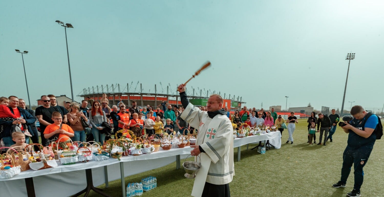 Święcenie pokarmów wielkanocnych na stadionie KGHM Zagłębie Lubin – ksiądz w ornacie błogosławi koszyczki z jajkami, babkami i chlebem, otoczone kibicami w barwach „Miedziowych” przy długim stole na murawie.
