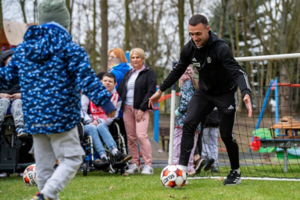 Piłkarz ŁKS Łódź w czarnym stroju sportowym prowadzi piłkę podczas zabawy z dziećmi na świeżym powietrzu w ramach akcji społecznej klubu.
