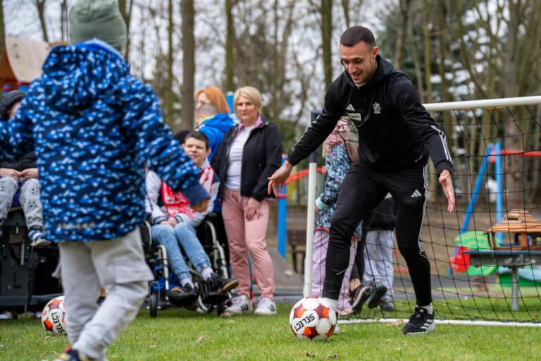 Piłkarz ŁKS Łódź w czarnym stroju sportowym prowadzi piłkę podczas zabawy z dziećmi na świeżym powietrzu w ramach akcji społecznej klubu.