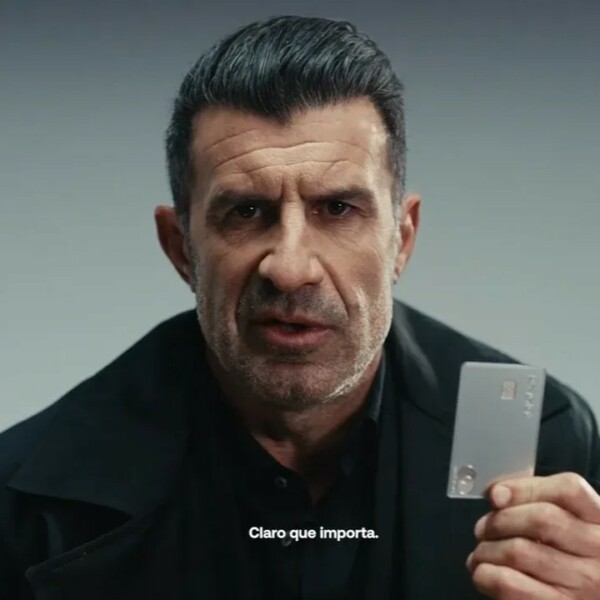 Luis FIgo w reklamie Revolut. Piłkarz trzyma srebrną kartę Revolut w lewej dłoni. Pozuje na tle szarej ściany i jest ubrany w czarną koszulę.