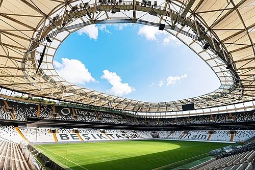Wnętrze stadionu Beşiktaş Park w Stambule z widokiem na murawę, czarno-białe trybuny i charakterystyczny otwarty dach linowy.